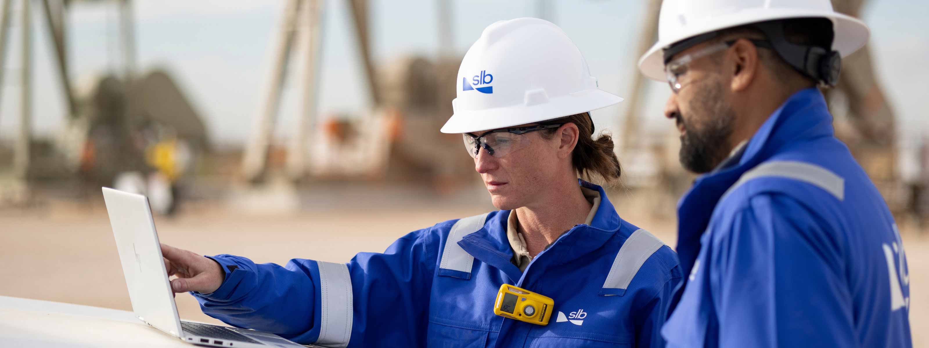 Two SLB field employees review information on a laptop at an oilfield site.